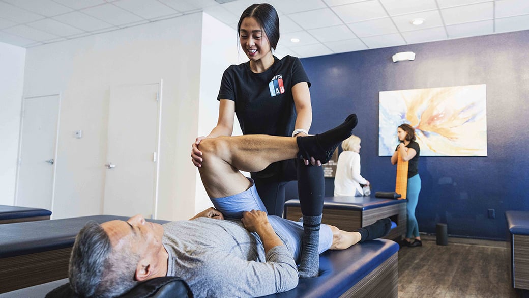 young middle aged man lying on padded stretch therapy bench with flexologist performing quad stretch on leg